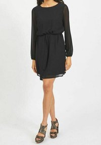 Robe noire en tissu léger avec des manches longues transparentes et une ceinture à nouer à la taille. Le modèle porte des chaussures à talons compensés rayées avec des découpes.