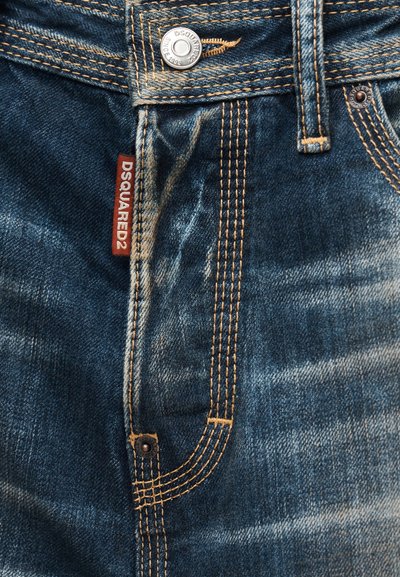 Κοντινό πλάνο μπλε τζιν με πορτοκαλί ραφές, μεταλλικά κουμπιά και κόκκινη ετικέτα ύφασμα που αναγράφει "DSQUARED2" στη ραφή της τσέπης.