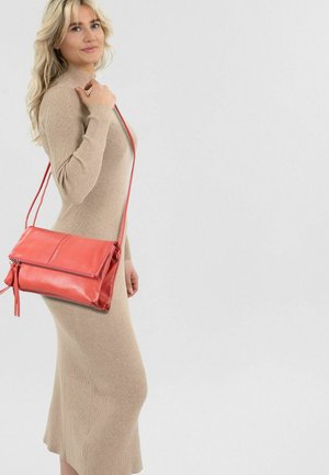Blonde Frau in einem beigen, langärmeligen, gerippten Kleid hält eine korallenrote Lederschultertasche mit Reißverschluss.