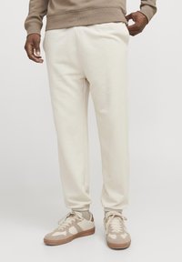 Crèmefarvede sweatpants med elastiske manchetter, sidelommer og en afslappet pasform, parret med beige sneakers med lyse accenter og teksturerede detaljer.