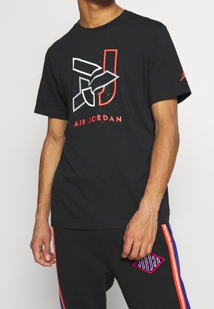 Mężczyzna noszący czarną koszulkę Air Jordan z czerwonym i białym logo oraz czarne spodnie z wielokolorowymi paskami i łatą Jordan.
