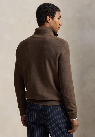 Άνδρας που φοράει καφέ πουλόβερ με ψηλό γιακά και ναυτικό μπλε παντελόνι με ρίγα, στέκεται πλαγίως σε ουδέτερο φόντο.