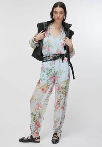 Femme portant une combinaison fleurie en tissu transparent avec des liens aux chevilles, une ceinture noire cloutée, un gilet en cuir noir, et des sandales noires cloutées.