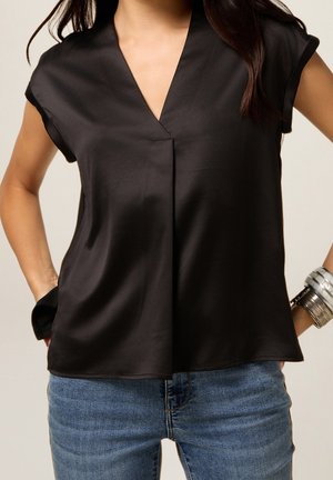 Kvinde iført en sort kortærmet V-hals bluse med en frontlægning, blå jeans, et sølvarmbånd og hænderne i lommerne.