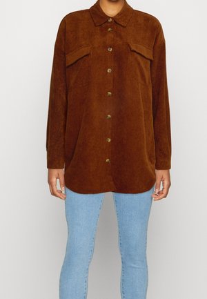 Brown corduroy button-up shirt with two flap chest pockets worn over light blue jeans, shown from mid-neck to thighs.