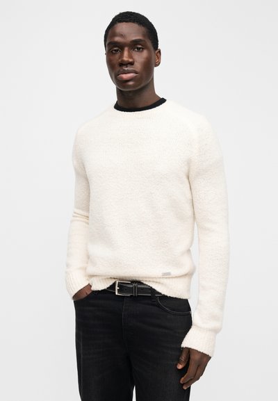 Light cream-colored sweater with a textured fabric and ribbed neckline, paired with dark jeans and a black belt.
