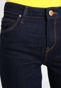 Mörkblå denimjeans med kopparstygn, featuring en knappstängning och en liten märkesetikett. Slät textur, klassisk five-pocket design.