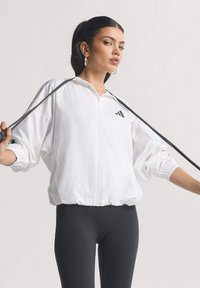 White zip-up jacket with elastic cuffs and hem, featuring a black logo on the left chest. Worn with black leggings, held by a model.