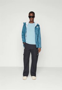 Jachetă ușoară de culoare teal cu accente albe, combinată cu o cămașă albastru deschis și pantaloni cargo negri. Modelul poartă ochelari de soare închiși la culoare și încălțăminte căptușită.