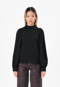Kvinne med Rett hår iført en svart langermet puffet bluse og lilla leopardmønstrede bukser, stående mot en hvit bakgrunn.