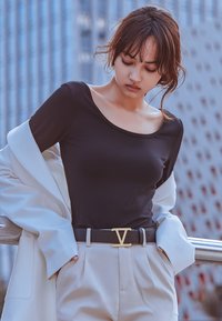 Zwarte lange mouw top met een ronde hals, gecombineerd met crèmekleurige high-waisted broek en een zwarte riem met een gouden embleem. Lichte jas over de arm gedrapeerd.