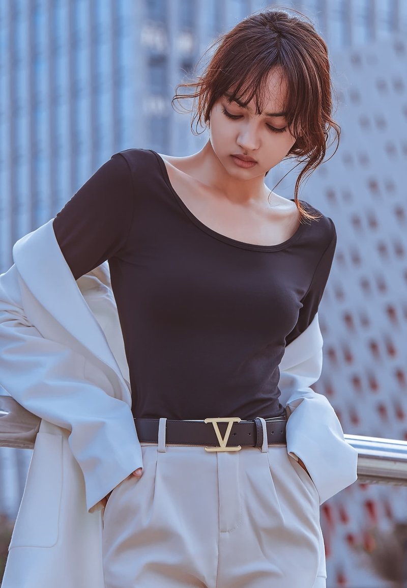 Zwarte lange mouw top met een ronde hals, gecombineerd met crèmekleurige high-waisted broek en een zwarte riem met een gouden embleem. Lichte jas over de arm gedrapeerd.