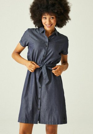 Vrouw met grote krullen die lacht, draagt een donkerblauwe korte mouwen jurk met knopen, gestrikt in de taille, tegen een effen achtergrond.
