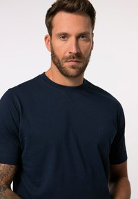 Marinblå t-shirt med kort ärm, tillverkad av mjuk bomull, med en rundad halsringning och en diskret logotyp tryckt på bröstet. Slät textur.