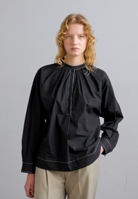 Camisa negra con un cuello alto, frente plisado y acentos de costura blanca. Mangas largas y un ajuste relajado.