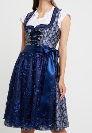 Woman wearing a blue and white traditional dress with lace blouse, patterned bodice, satin waist tie, and floral embroidered apron.