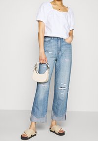Top corto blanco con mangas abullonadas, jeans rasgados de color azul claro, un pequeño bolso de crema y sandalias deslizantes beige. Conjunto informal con un ajuste relajado.