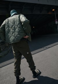 Veste olive verte matelassée, pantalon cargo avec poches et sandales sombres. Textures mates et vêtements superposés dans un cadre urbain.