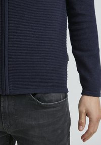 Námornícky pletený kardigán s horizontálnym ryhovaním, so zipsom vpredu, úzkym strihom a kontrastnými manžetami, skombinovaný s tmavými džínsy.