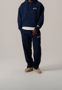 Marine hoodie met een voorzak en witte tekst, gecombineerd met bijpassende broek. Tan schoenen versterken de casual uitstraling met textuurstof.