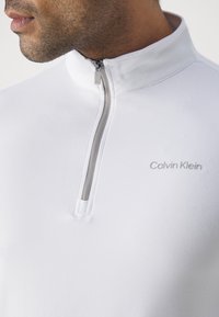 Primer plano de un hombre que lleva una sudadera blanca de Calvin Klein con cremallera, de cuello alto y con el tirador de la cremallera visible marcado con "CK."
