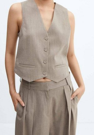 Beżowa kamizelka bez rękawów w cienkie prążki na cztery guziki, zestawiona z dopasowanymi, plisowanymi, wysokim stanem spodniami, ręce w kieszeniach.