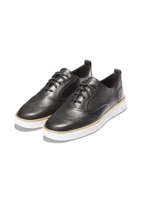 Un paio di scarpe in pelle nera stile brogue con suole in gomma bianca, lacci e perforazioni decorative sulla parte superiore.