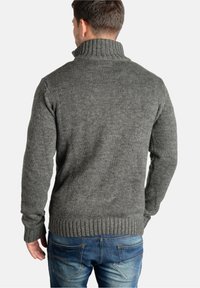 Grijze gebreide trui met een hoge geribbelde kraag en lange mouwen, voorzien van een textuurpatroon en een aangesloten tailleband. Draagt blauw spijkerbroek.