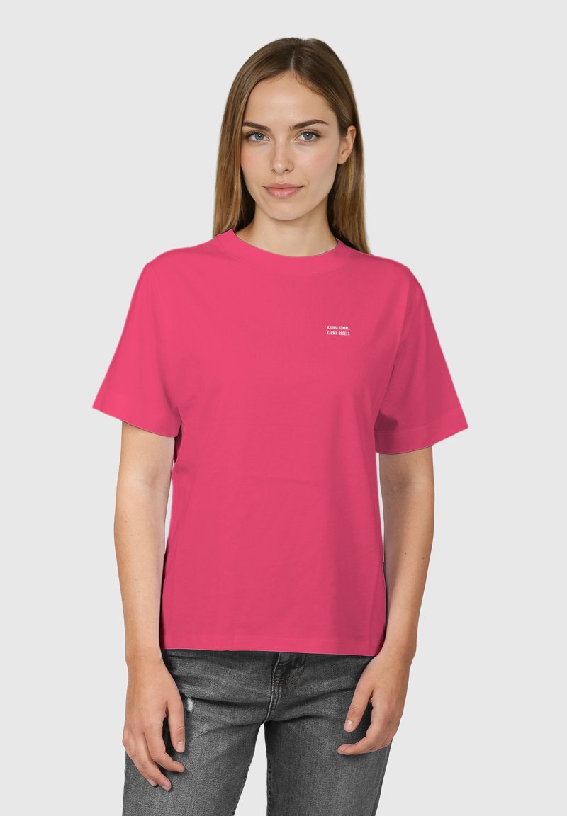 Femme portant un t-shirt à manches courtes rose vif avec un petit texte blanc sur la poitrine et un jean noir délavé, debout devant un fond uni.