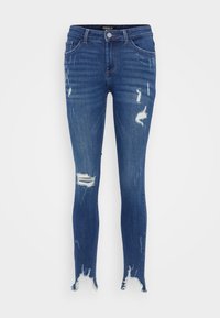 Sötétkék, elnyűtt hatású, skinny farmernadrág, amelyen több szakadás és kavart szárvégek találhatók, valamint közepes magasságú derékpánt.