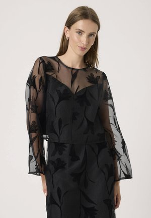 Fiatal nő fekete, áttetsző virágos hosszú ujjú felsőt visel, amelyhez fekete, virágos nadrág tartozik, egyszerű világos háttér előtt áll.
