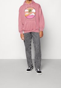 Sudadera con capucha rosa con un gráfico frontal que presenta montañas y un sol, combinada con jeans de denim gris y zapatillas negras.