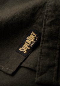 Donkergroen stof met een gestructureerde weefsel, met een zwart label waarop "SUPERDRY" in gouden letters staat. De stiksels zijn zichtbaar langs de randen.