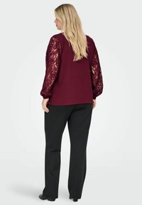 Top bordeaux con tessuto strutturato e maniche a sbuffo in pizzo trasparente, abbinato a pantaloni neri. Il pizzo presenta motivi floreali e dettagli a coste.