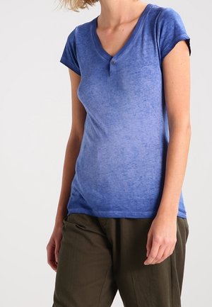 Mujer vistiendo una camisa henley azul ajustada de manga corta y pantalones sueltos de color verde oliva, de pie contra un fondo claro y liso.