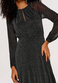 Musta, kimaltava mekko, jossa on läpikuultavat pitkät hihat, avainreikäkaulus ja kerätty vyötärö, joka luo virtaavan siluetin.