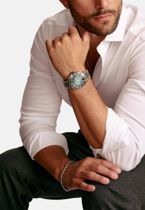Silver metal watch with a green dial and multiple dials, worn on a wrist. White long-sleeve shirt, gray pants, and a silver chain bracelet visible.