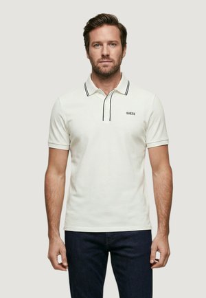 Homme adulte aux cheveux courts bruns et à la barbe, portant un polo blanc à manches courtes avec bordure noire et un jean bleu foncé, debout de face.