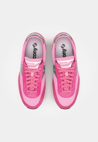 Rozā sporta apavi, kas izgatavoti no gludām un strukturētām materiālēm, ar zaļa akcenta elementiem, mīkstu zoli un tonāliem šņorēm. Redzams Saucony logo.