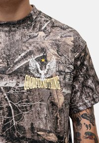 T-shirt w wzór moro w odcieniach brązu i zieleni. Posiada białą grafikę skrzydeł oraz tekst „GOODFORNOTHING” w kolorze żółtym. Krótkie rękawy.