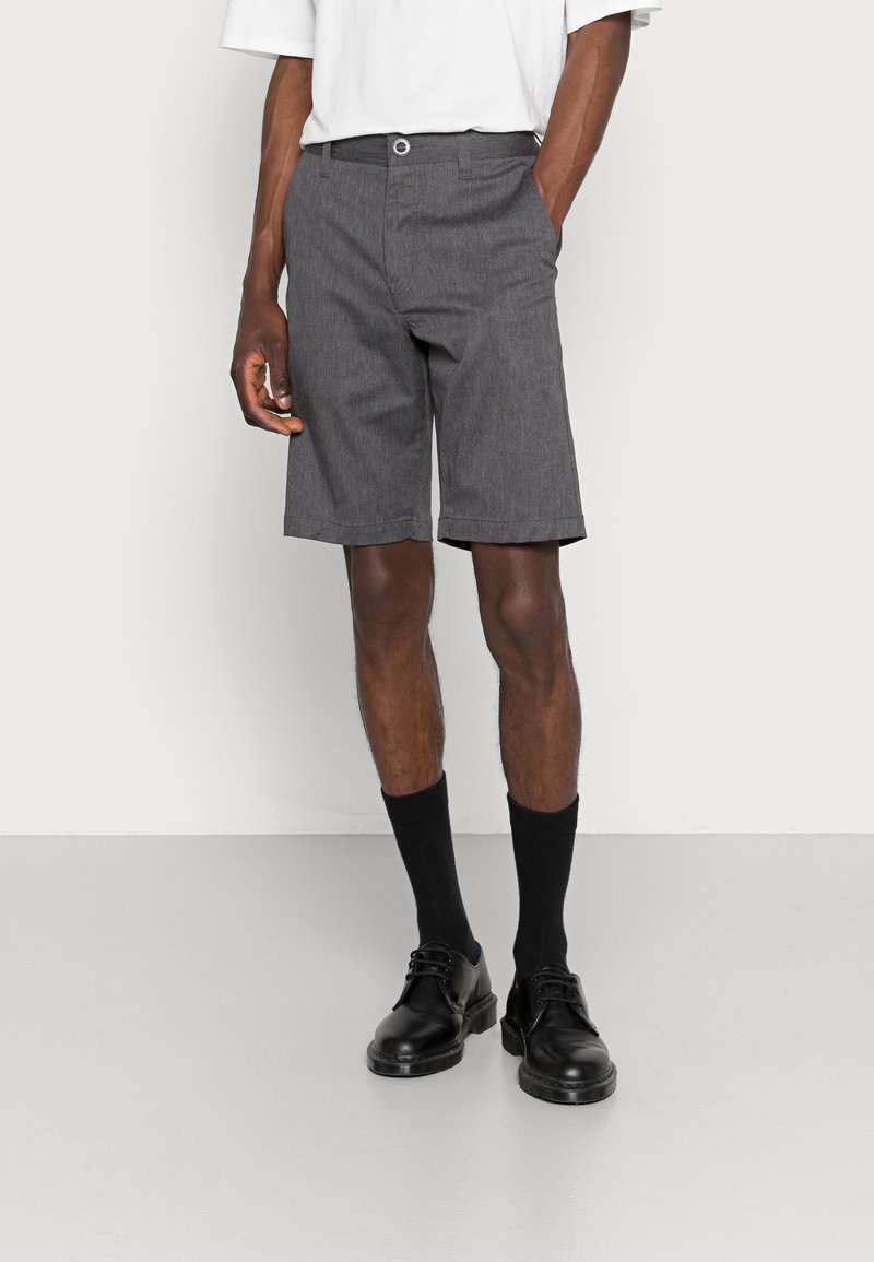 Shorts gris en tissu texturé, dotés d'une fermeture à bouton, de poches latérales et d'une coupe arrivant au genou. Portés avec des chaussettes noires et des chaussures.