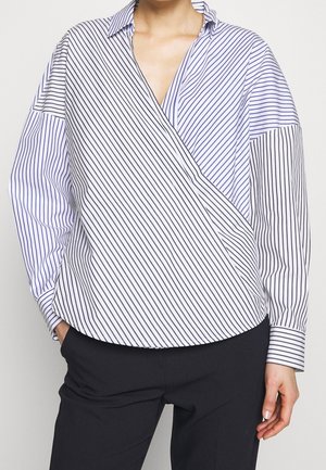 Femme portant un chemisier blanc à manches longues avec de fines rayures diagonales et verticales noires et bleues, associé à un pantalon noir.