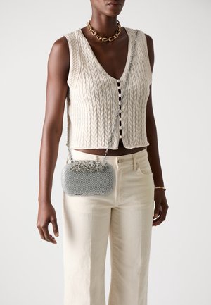 Femme noire portant un gilet court crème sans manches en maille torsadée, un pantalon crème taille haute, un collier chaîne doré et un sac bandoulière argenté texturé.