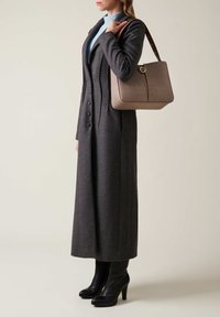 Frau in einem langen dunkelgrauen Mantel und schwarzen High Heels, die eine beigefarbene strukturierte Handtasche über der Schulter trägt, vor einem einfarbigen Hintergrund.