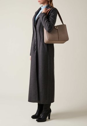 Frau in einem langen dunkelgrauen Mantel und schwarzen High Heels, die eine beigefarbene strukturierte Handtasche über der Schulter trägt, vor einem einfarbigen Hintergrund.