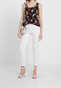 Top sin mangas con patrón floral en azul marino, combinado con jeans blancos ajustados y tacones beige con tiras. Pequeño bolso con acentos en contraste.