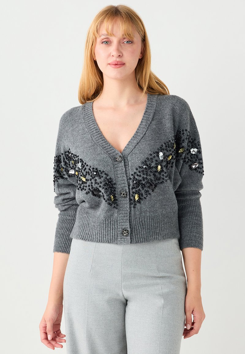 Cardigan gris avec col en V, boutons sur le devant, poignets côtelés et un motif décoratif en perles noires et transparentes.