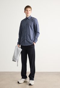 Striped button-up skjorte i marineblå, kombinert med mørke bukser. Personen holder en lys grå hettegenser. Uformelle sko har en kraftig design.