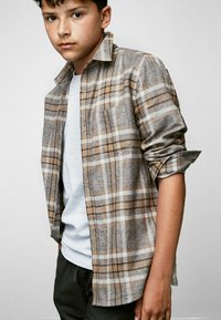 Rutig flanellskjorta i brunt och beige, med en grå t-shirt under. Skjortan har upprullade ärmar och en enkel bröstficka.