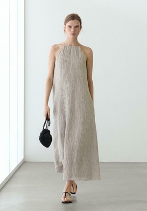 Kobieta w bezrękawnikowej, teksturowanej beżowej sukni maxi, niosąca małą czarną plecioną torebkę, spacerująca po minimalistycznym białym pokoju.